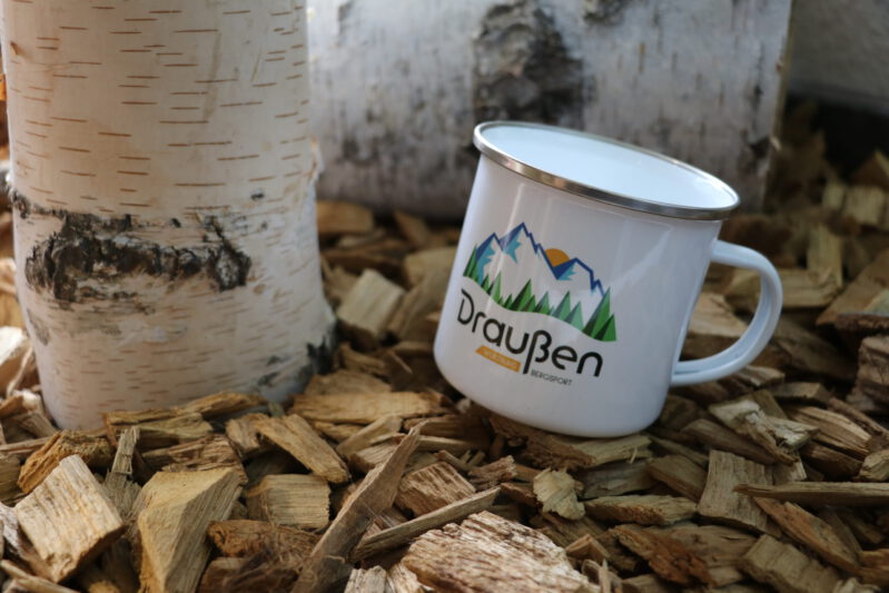 Tasse mit Draußen-Logo auf Rindenmulch neben einem Baumstamm